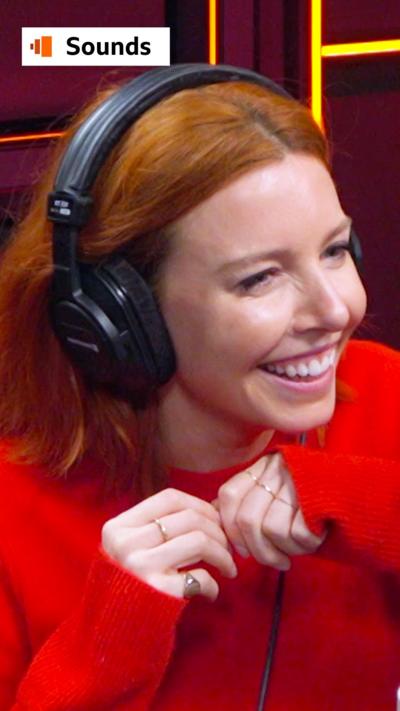 Stacey Dooley smiles in a radio studio - she has red hair and wears a red jumper and black headphones 