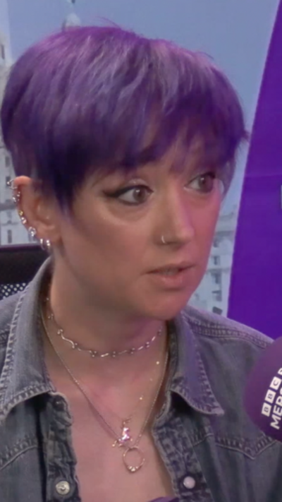 Person with short purple hair speaking into a microphone labeled “BBC Radio Merseyside,” seated indoors with a purple BBC backdrop visible