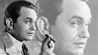 Black and white image: Edward G Robinson on the left hand side of the image, looking towards the camera through the corner of his eye whilst smoking a cigar. On the right hand side is a faded, enlarged version of his head from the same image, which is in the background.