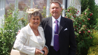 Sylvia Tadd on the left wearing a white top and jacket with a cream fascinator. Bob is on the right and he has his arm around Sylvia. He is wearing a black suit with a white shirt and a purple tie.