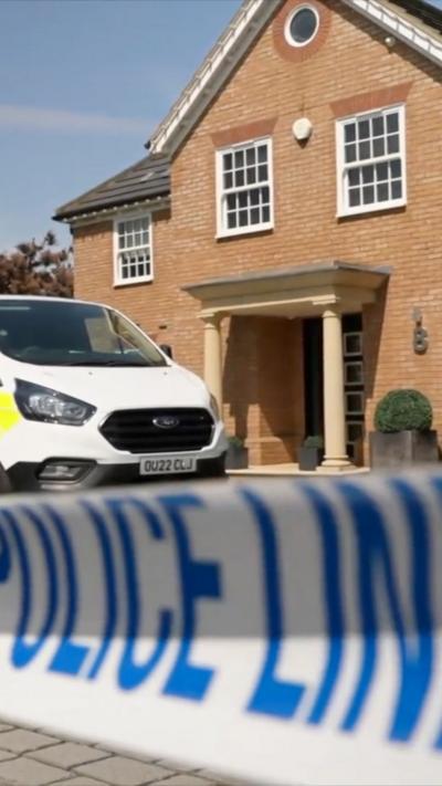 A police cordon is in focus while the front of a white police van comes into frame with a large, orange brick house in the background with a pillar porch