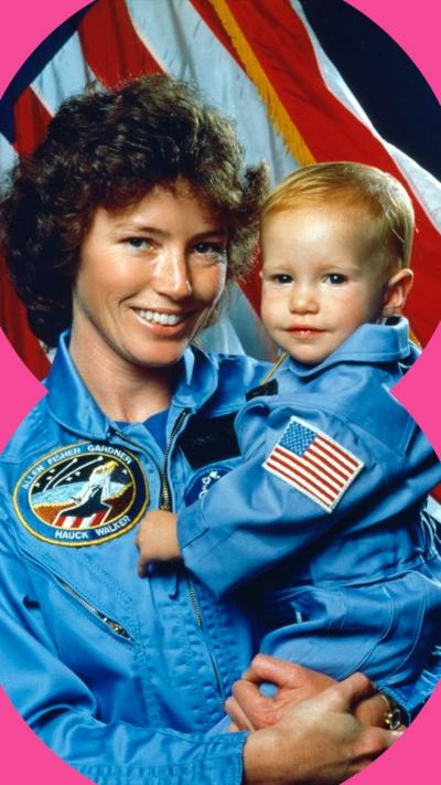 Archive photo of Anna Fisher and her baby daughter, both in blue Nasa uniforms, pose in front of the American flag