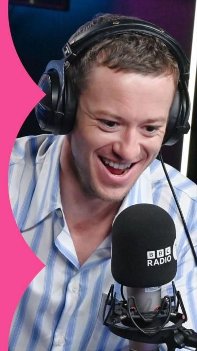 Joseph Quinn wearing a stripy shirt in the R1 studio in front of a mic and wearing headphones.