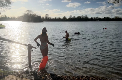 A group of swimmers walk into South Cerney lake on a clear bright winter's day. The sun is reflecting off the water and the trees that border the lake can be seen in the distance