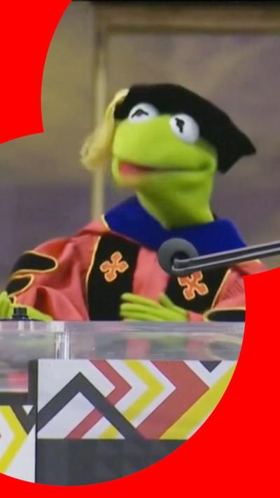 Kermit stands in cap and gown behind a lecturn