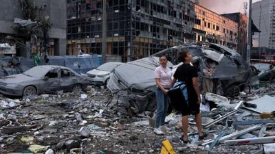 Man and woman stand in rubble looking up with destroyed cars around them holding hands