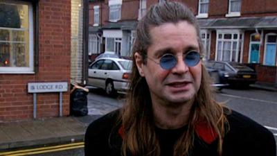 Ozzy Osbourne stands on the street where he grew up, he is dressed in black and wearing blue tinted glasses. Behind him is the road sign, Lodge Road, and you can see terraced homes. 