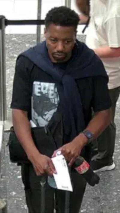  A man passes through an airport scanner holding a white envelope and a Coca‑Cola bottle, wearing a dark T‑shirt, a sweater over their shoulders, and a smartwatch, with a shoulder bag at their side and other travellers in the background.