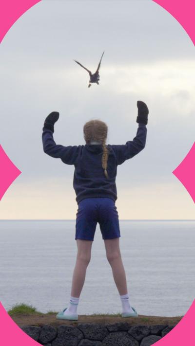 A young girl stands on a cliff edge with her arms raised watching a puffin fly off over the sea