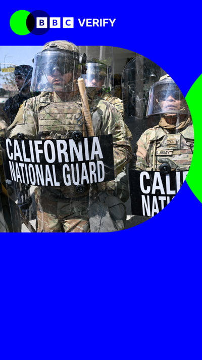 National Guard troops stand in a row. They are wearing military gear and hold riot shield with California National Guard written on them. The troops also wear helmets with visors on them. 