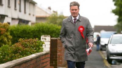Keir Starmer walking on a residential street towards the camera in 2017. He is wearing a red labour rosette and a rain mac