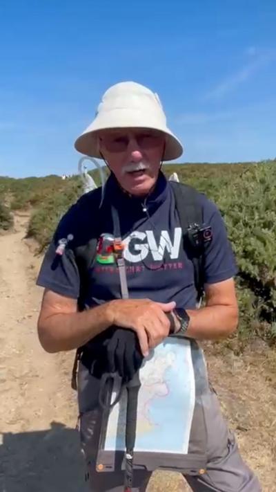John Erryl Parry, who is stood outside and is wearing a white bucket hat. He has a grey moustache and glasses. He is holding a map and has his arms crossed.