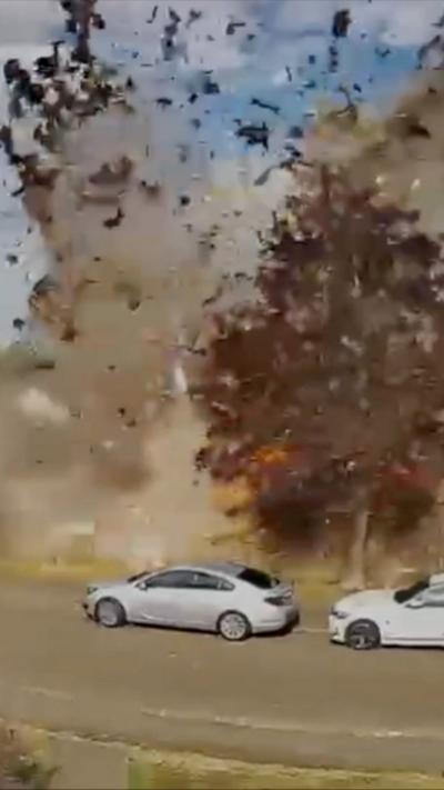 A plume of smoke and debris erupts beside a roadside, rising high into the air from a powerful explosion. Two silver cars sit close to the blast, partially lit by the bright flash. A tree with reddish‑brown leaves stands near the explosion.