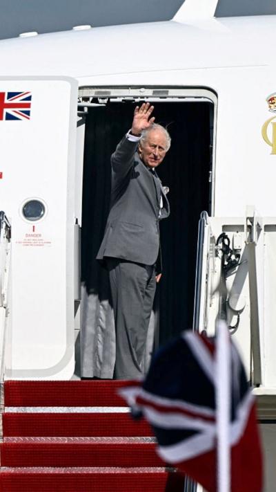King Charles stands in the doorway of a white airplane with the British flag on it as he waves.