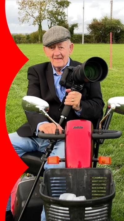 Mo Hunt holds his camera, sat with a camera in a mobility scooter on a rugby pitch