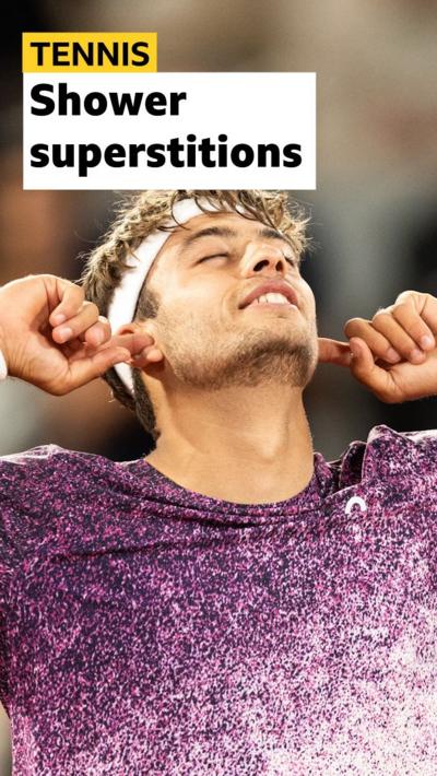 Flavio Cobolli celebrating his French Open win by putting his fingers in his ears. He is wearing a white headband and a purple shirt.