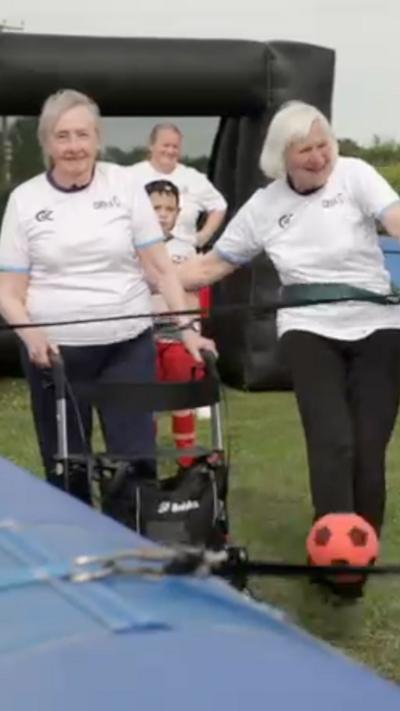 Two women in white t-shirts playing football with an orange and black ball. they are attached to bungee cords