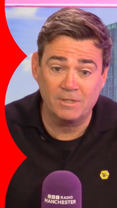 A man, Andy Burnham, has short brown hair and wears a black top. He's sitting in front of a purple Radio Manchester microphone