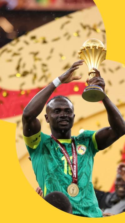 A Senegalese footballer wears a green football shirt and a gold medal. He is holding up the Africa Cup of Nations trophy