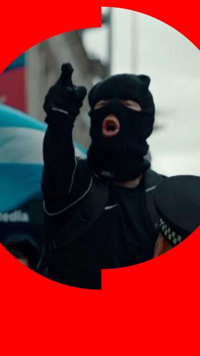 Person in balaclava pointing while shouting as a police officer watches