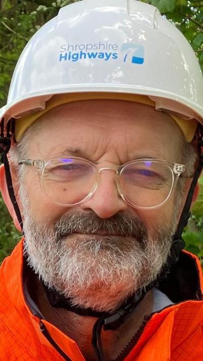 A man wearing a hard hat which has Shropshire Highways written on it. He is wearing glasses, has a beard and a high vis jacket