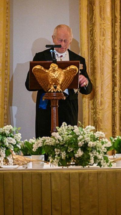 King Charles III, standing behind a podium with a gold eagle, addresses guests in a ballroom with gold curtains, gold table cloths and greenery on the tables.