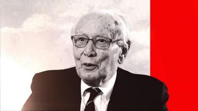 An elderly man wears glasses, a black jacket and a black tie