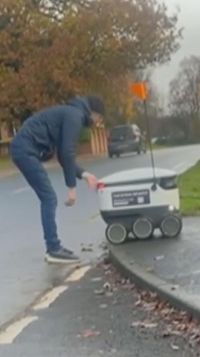 Man bends over to push white delivery robot onto pavement.