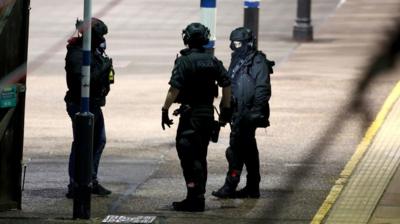 Three specially trained officers dressed in dark clothing, helmets and gloves stand on a train station platform