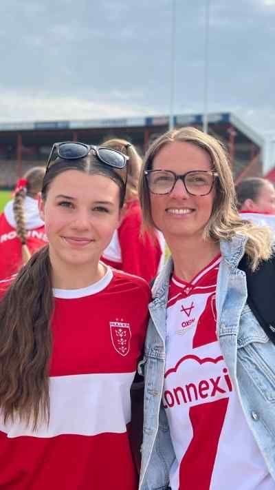 Two Hull KR fans smiling for a photo at a rugby ground, wearing red and white team shirts, with other supporters and stadium seating in the background.