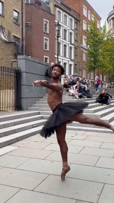 A male ballet dancer wearing a black tutu dances on the streets of London while others watch behind him
