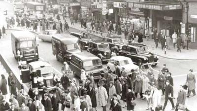 Large number of pedestrians cross the street while several cars are lined up