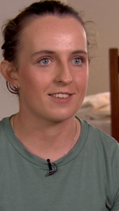 A young woman, Amy Symington, smiles broadly at the camera. She is sitting in the living room of a home. In the background, through an entrance into another room we can see a dining table. There is a also a low bookcase filled with books and topped with family photos. Amy has dark hair tied back and is wearing a light green t-shirt.