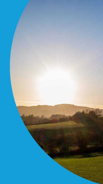 The sun sets in a blue sky. It is starting to disappear behind a hill. Its rays are illumination several fields in the foreground.