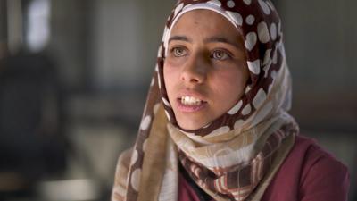 A young woman wearing a head scarf sits.