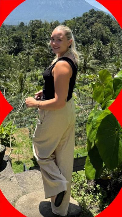 A woman wearing a back sleeveless top and beige trousers stands with a forest in the background