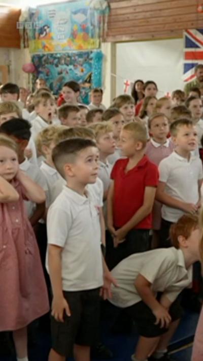 Dozens of schoolchildren in a school hall. the boys and girls are sat on benches and wearing an assortment of white polo shirts, red tops and pink dresses