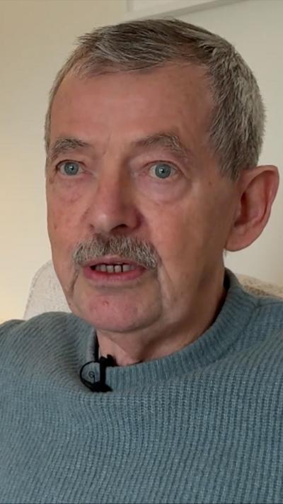 Elderly man with grey hair and moustached wearing blue jumper indoors