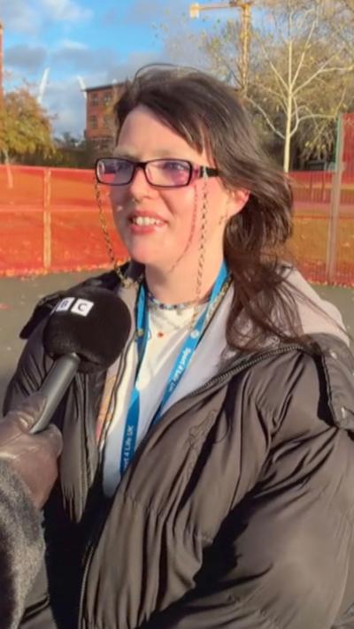A young woman with brown hair wears glasses with a chain on while she speaks into a black BBC microphone