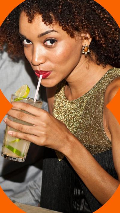 A woman looking to the camera, sipping from a white straw, holding a drink with lime wedges. She wears a gold top and red lipstick.