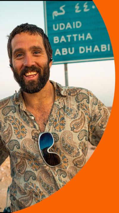 A man, Mithcell Hutchcraft wears a patterned shirt. Behind him is a road sign pointing to Abu Dhabi