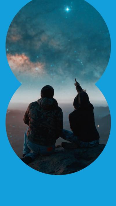 Two people sat on a mountain top looking at the night sky. One points to the sky.