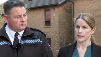 A male police officer with short grey hair in uniform, stood next to a woman with tied-back blonde hair, wearing a green blouse and black suit jacket. They are stood in front of a brown block of flats.