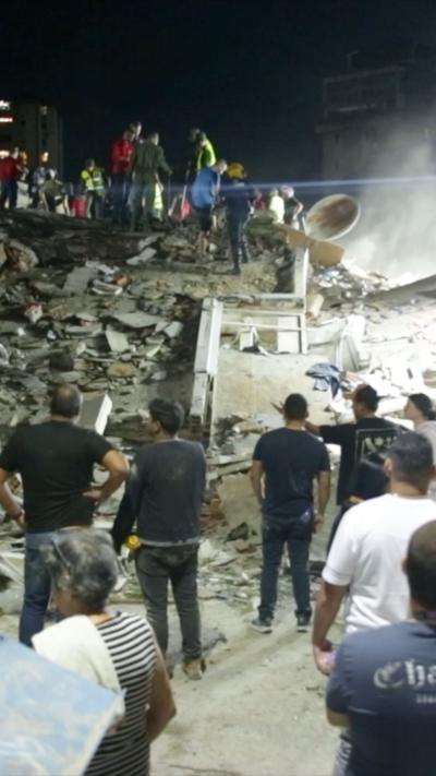 A number of people stand in front of a collapsed building which is a large pile of rubble with a number of rescuers standing on top