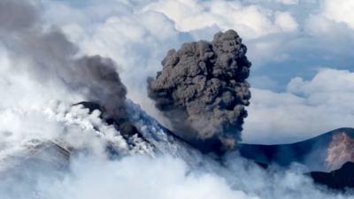 A black plume erupts from Mt Etna with clouds behind it.