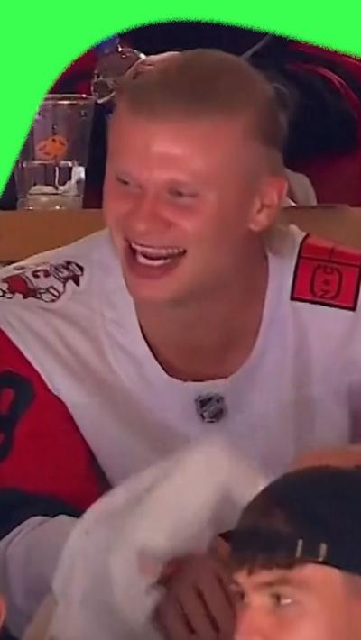 Norway football player Erling Haaland, laughs as he watches an ice hockey match from the stands. He's wearing a white ice hockey jersey.