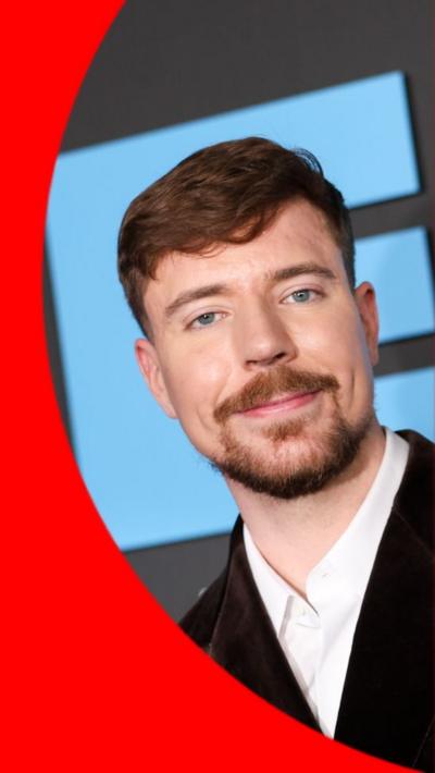 MrBeast aka Jimmy Donaldson has light brown hair, a beard and moustache. He is wearing a dark-coloured velvet suit jacket.