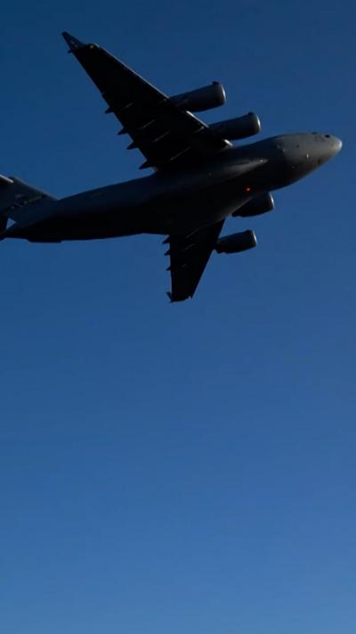 US military plane flies overhead, it is dark grey against a blue sky