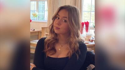 An image of Jessica Glasser sitting at a table. She is wearing a long sleeve black top and gold necklaces. She has long, waved blonde hair and is smiling at the camera.