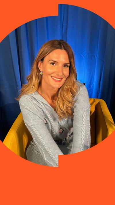 A woman sits in a yellow chair in front of a blue curtain. She has long hair and is wearing a grey top.